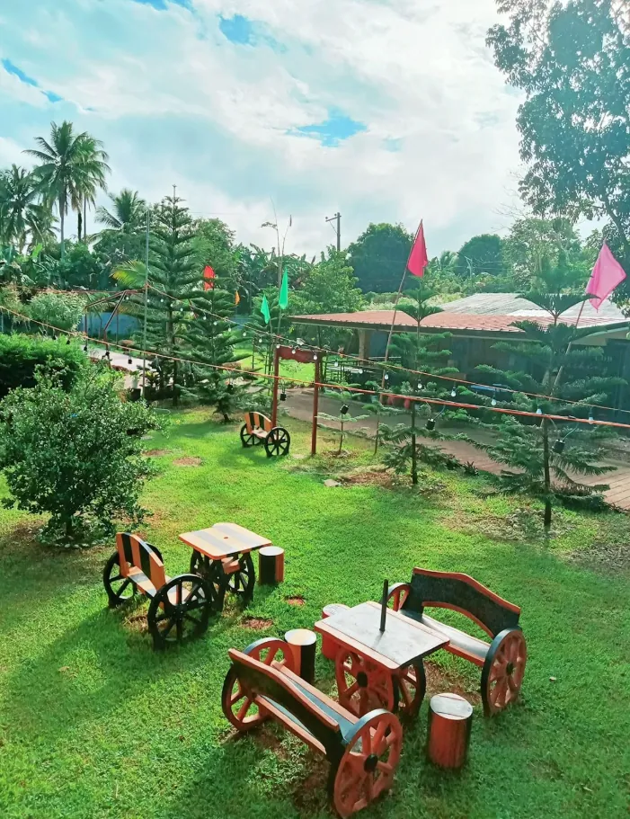 View over the camp's garden with pine trees, wooden cart-wheel benches and festive flags under a blue sky