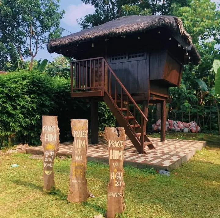 A wooden prayer kubo on stilts beside carved posts reading Praise Him
